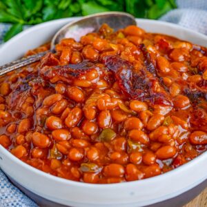 Square image of Homemade Baked Beans in bowl with spoon and bacon showing.