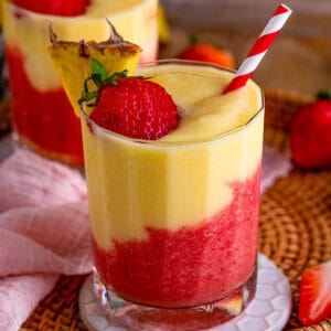 Square image of one drink close up layered and garnished with pineapple, strawberry and straw.
