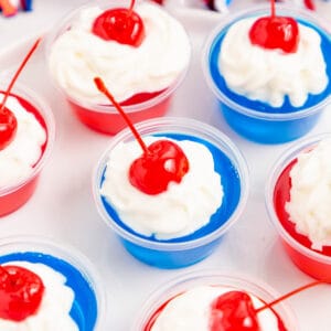 Square image close up of jello shots on white plate topped with whipped cream and cherries.