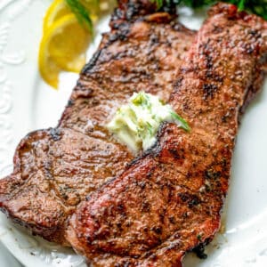 Square image of two steaks cooked on white plate with butter.