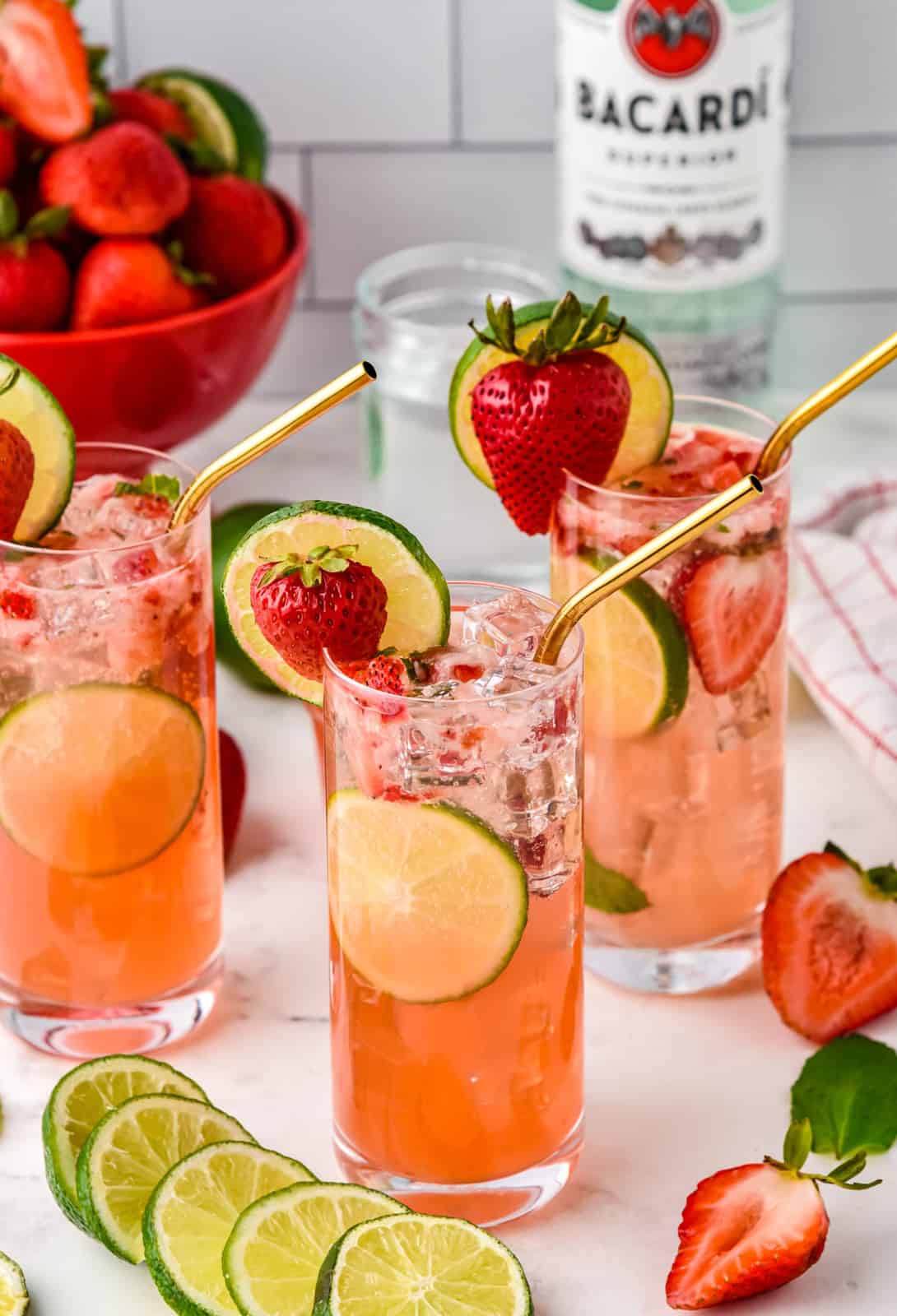 Three Strawberry Mojitos with strawberries and alcohol in background.