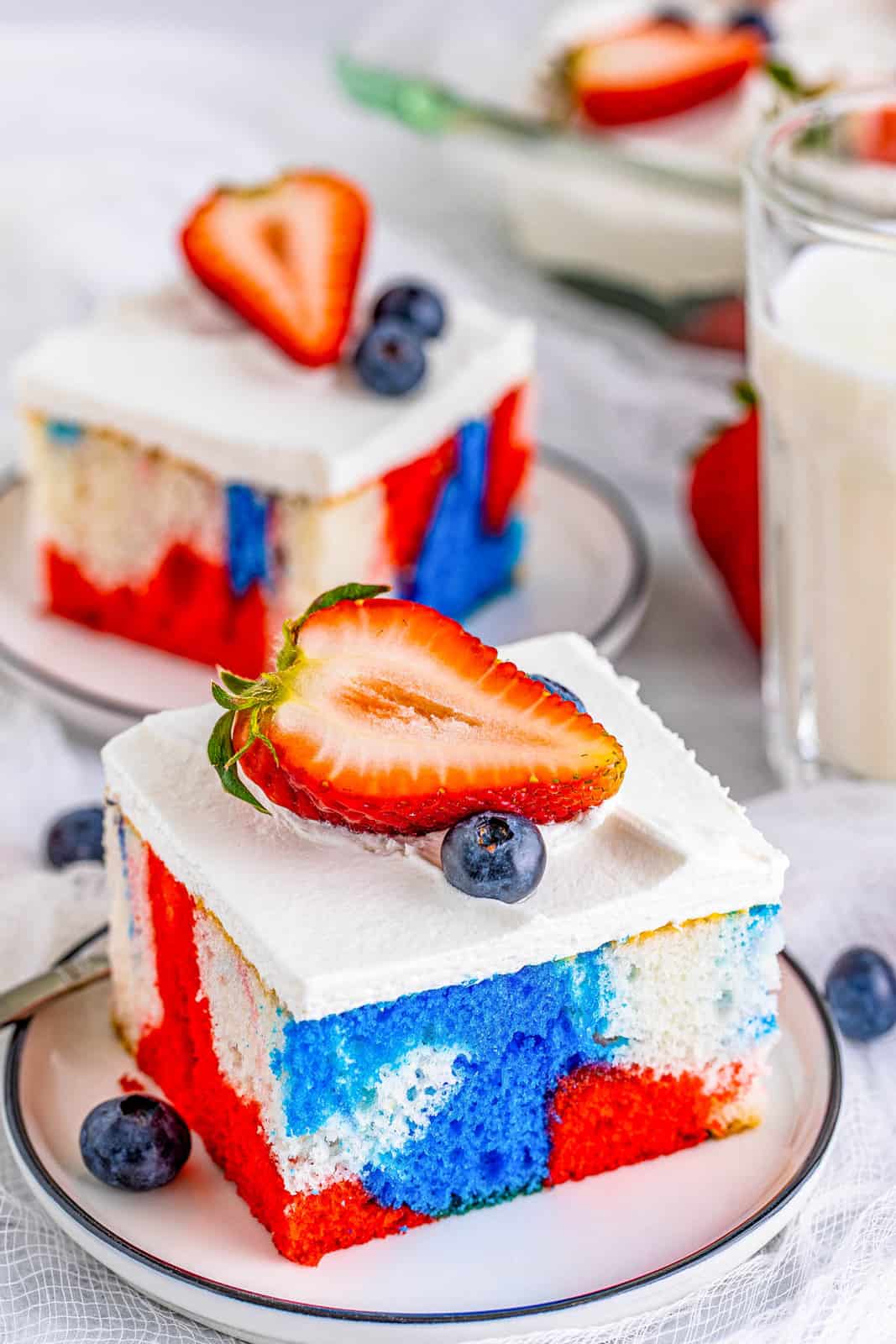 Two slices of Tie Dye 4th of July Cake on white plates topped with berries.