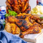 Square image of pieces of chicken on white board with whole chicken and beer in background.