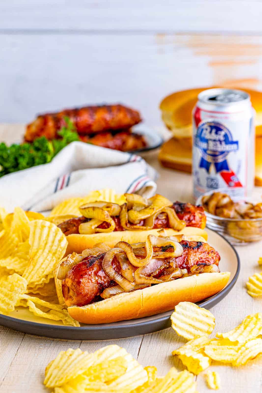 Cheese Stuffed Bacon Wrapped Brats on plate with beer and chips in back and around plate.