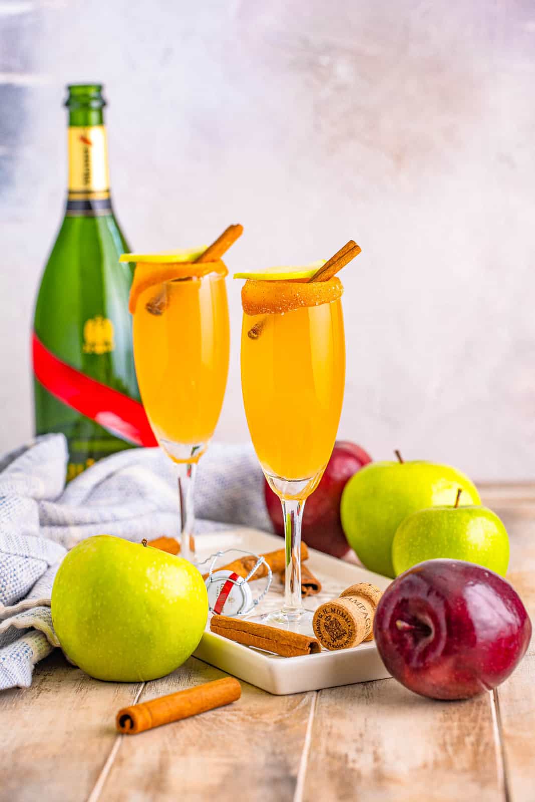 Two Apple Cider Mimosas with garnishes on white platter with apples and champagne.