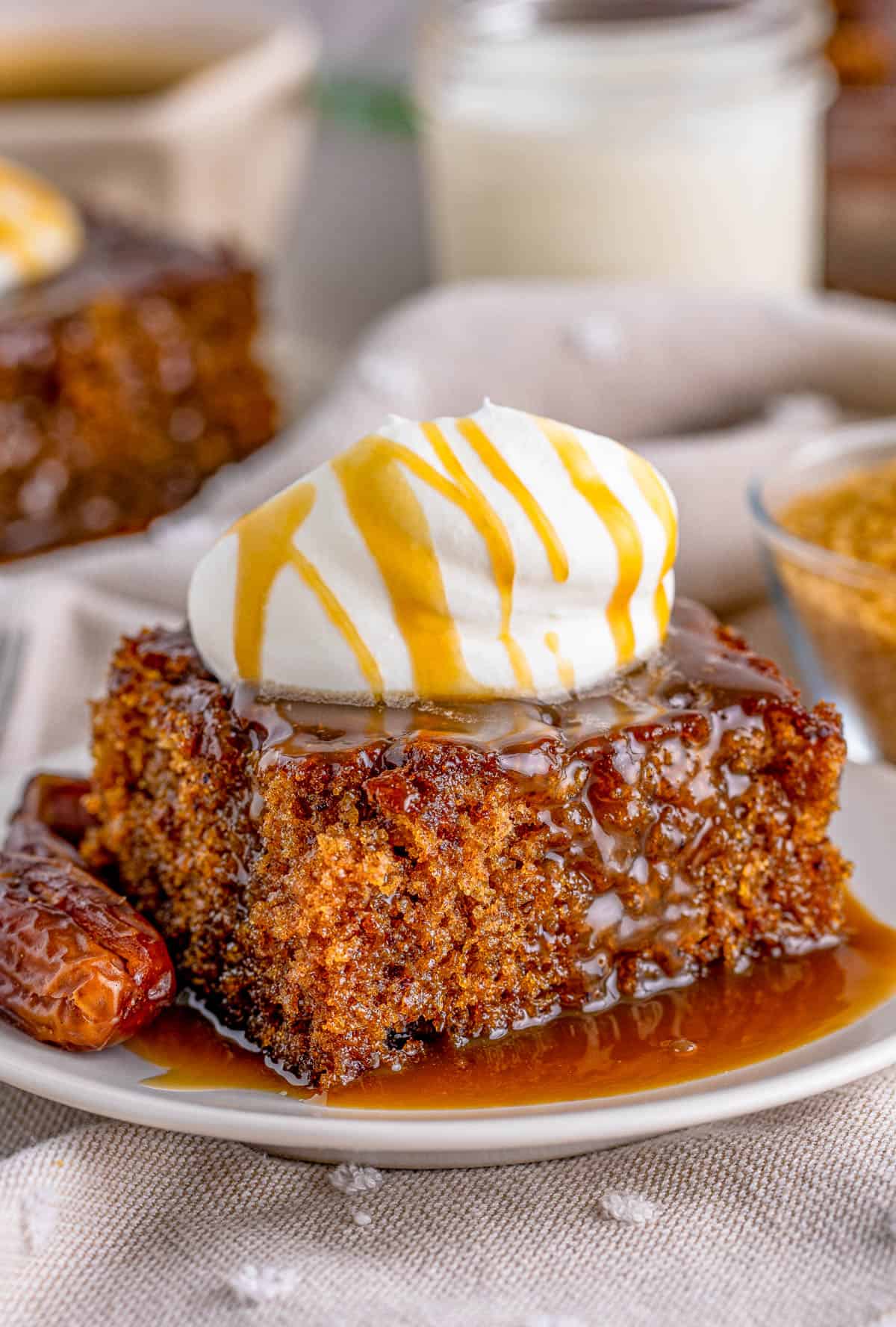 Slice of the Sticky Toffee Pudding Recipe on plate with dates, whipped topping and more sauce poured over top.