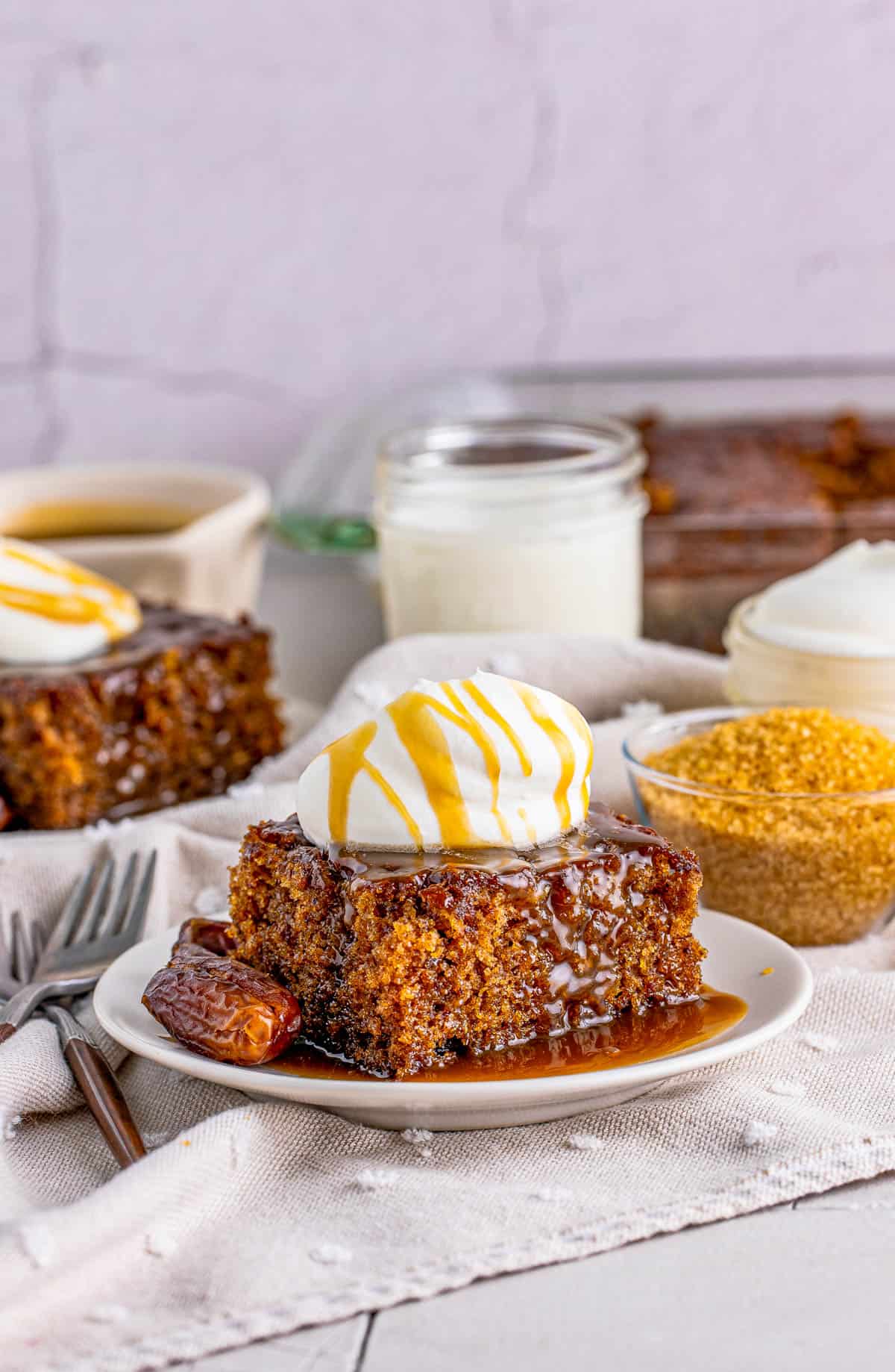 Sticky Toffee Pudding Recipe on white plates with whipped cream on top, milk and sugar in background.