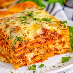 Close up square image of a slice of lasagna on white plate topped with garnishes.