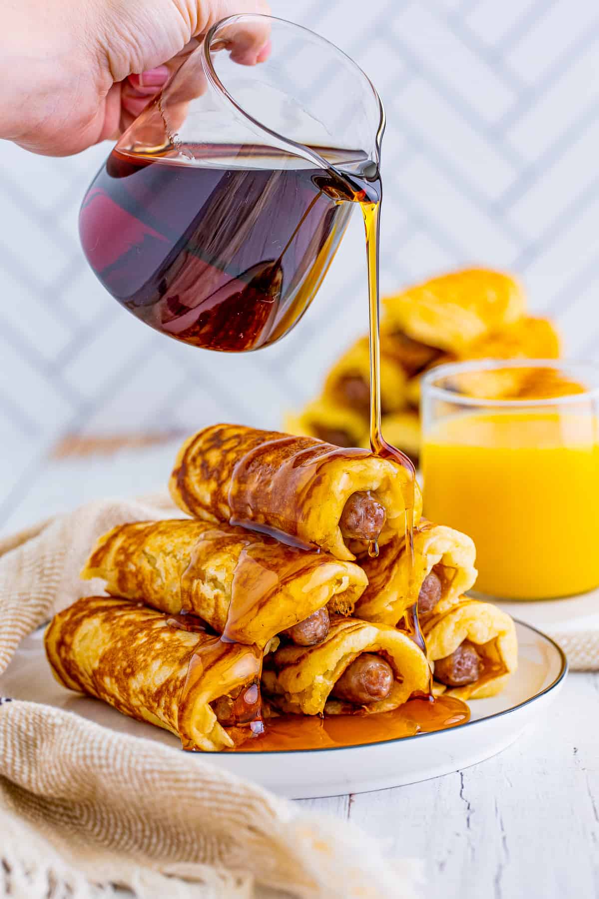 Syrup being poured over a stack of Breakfast Pigs in a Blanket on white plate.