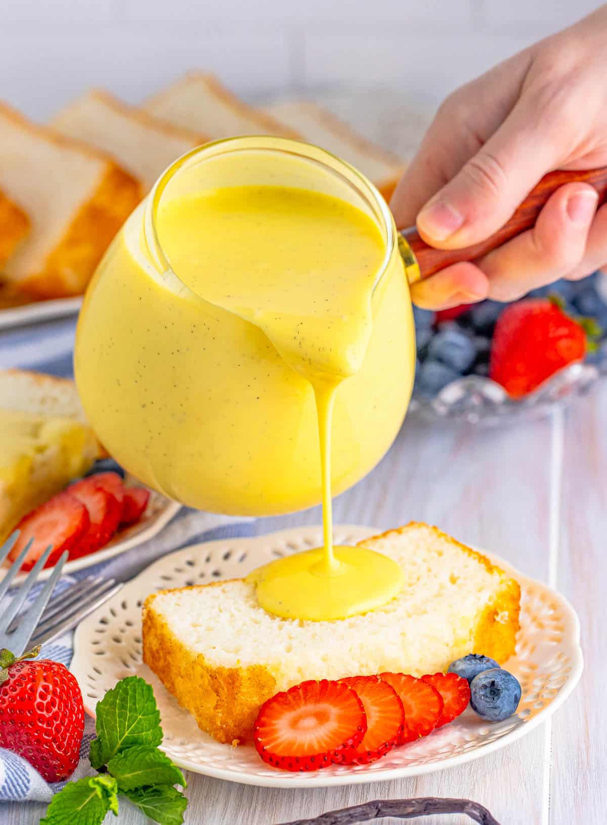 Crème Anglaise being poured over a slice of angel food cake on plate with fruit.
