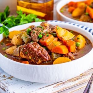 Square image of Beef Stew in white bowl with spoon next to it and parsley in background.