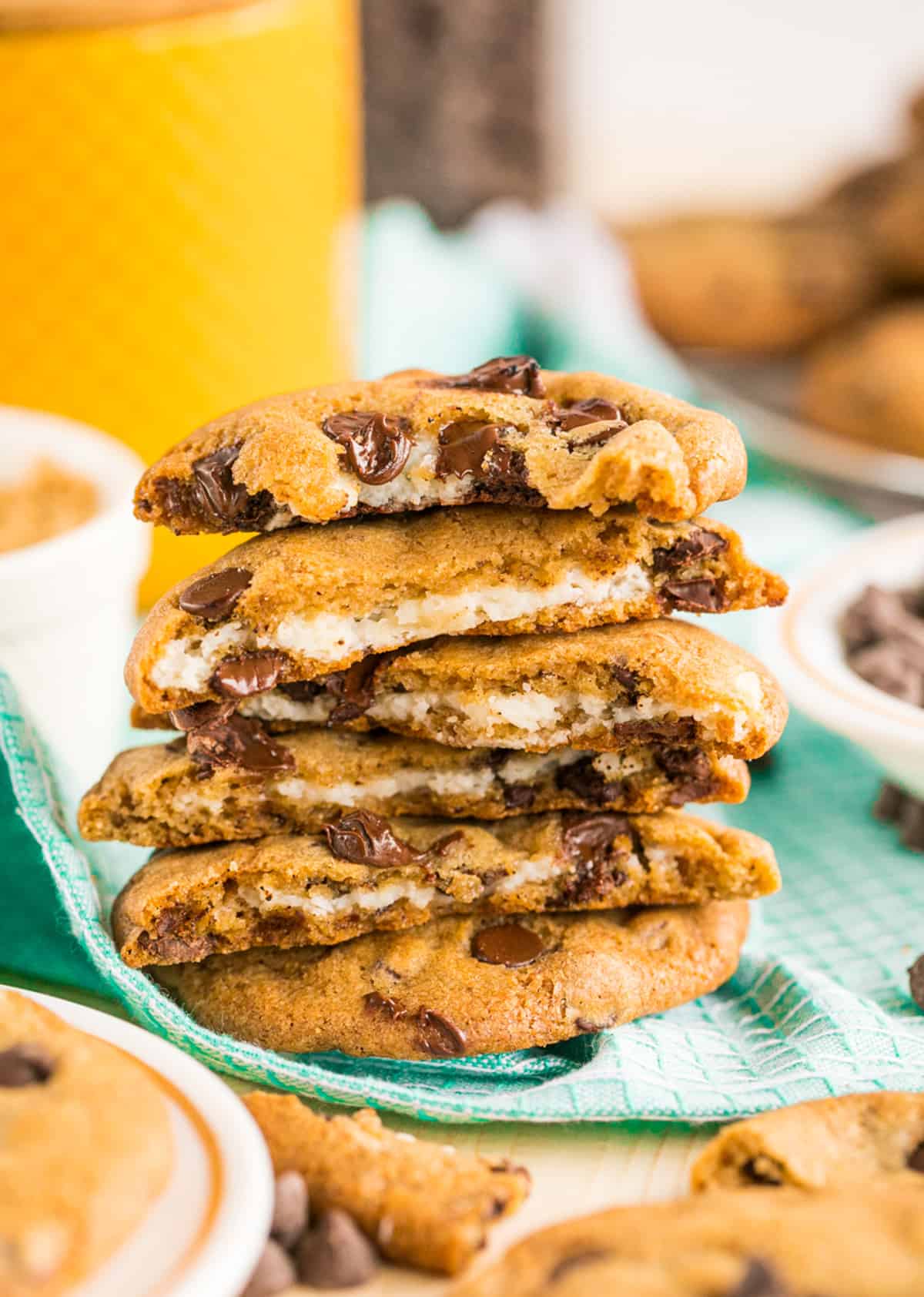 Stacked Cheesecake Stuffed Chocolate Chip Cookies on teal linen with whole cookies around it split in half showing cheesecake filling.