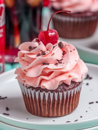 Square image of one cupcake frosted on plate with cherry and chocolate shavings on top.