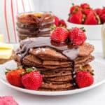 Square image of pancakes with chocolate sauce drizzling down the side topped with strawberries.