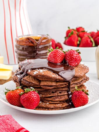 Chocolate Pancakes on white plate with powdered sugar, chocolate sauce and topped with strawberries.