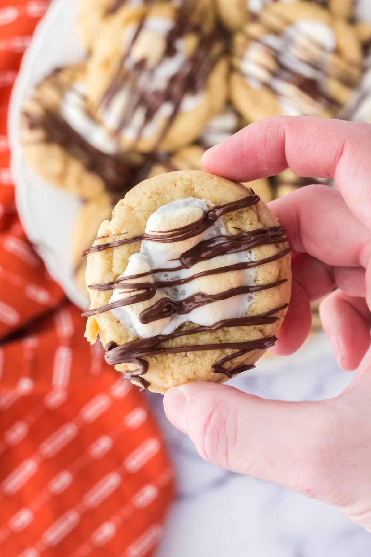 Hand holding up one finished cookie showing melted marshmallow and chocolate drizzle.