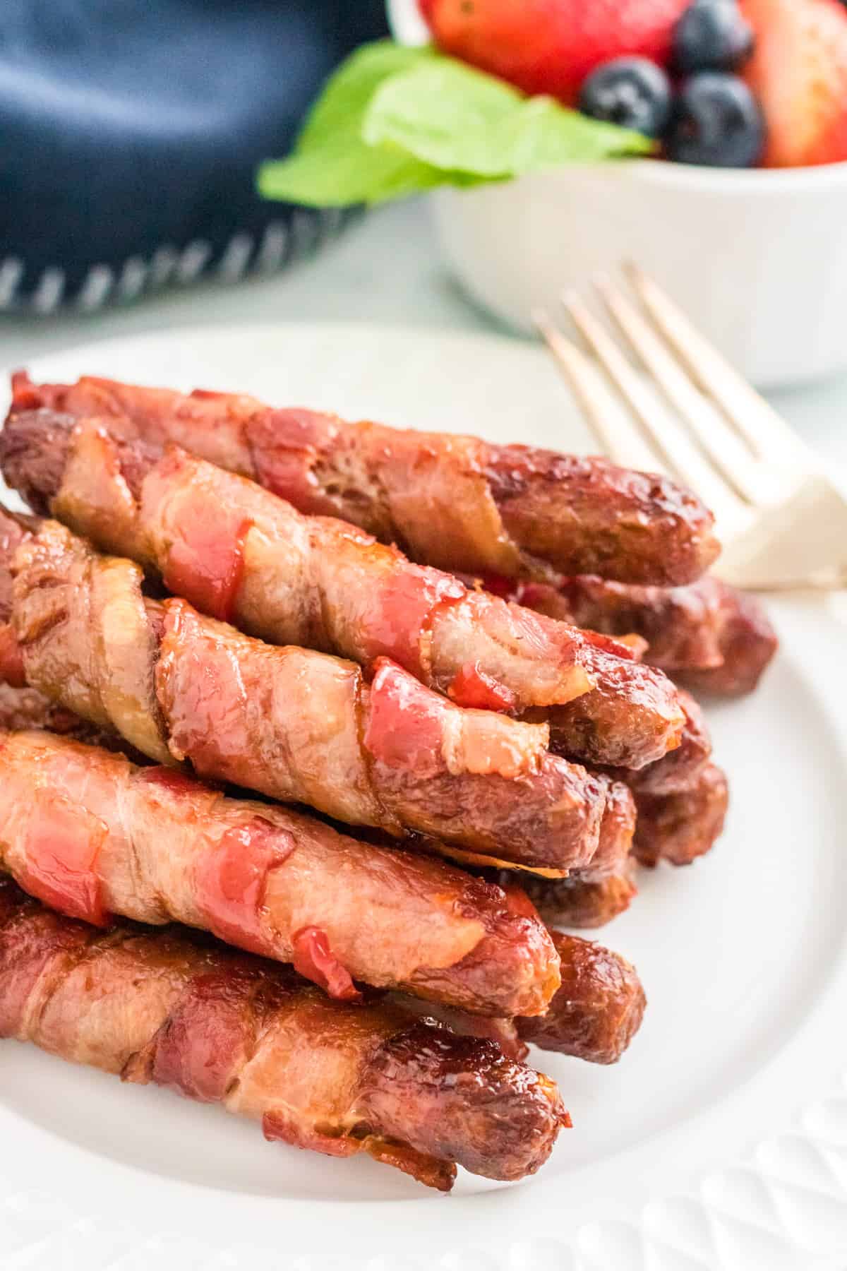 Bacon Wrapped Sausages stacked on white plate with fork and bowl of fruit in background.