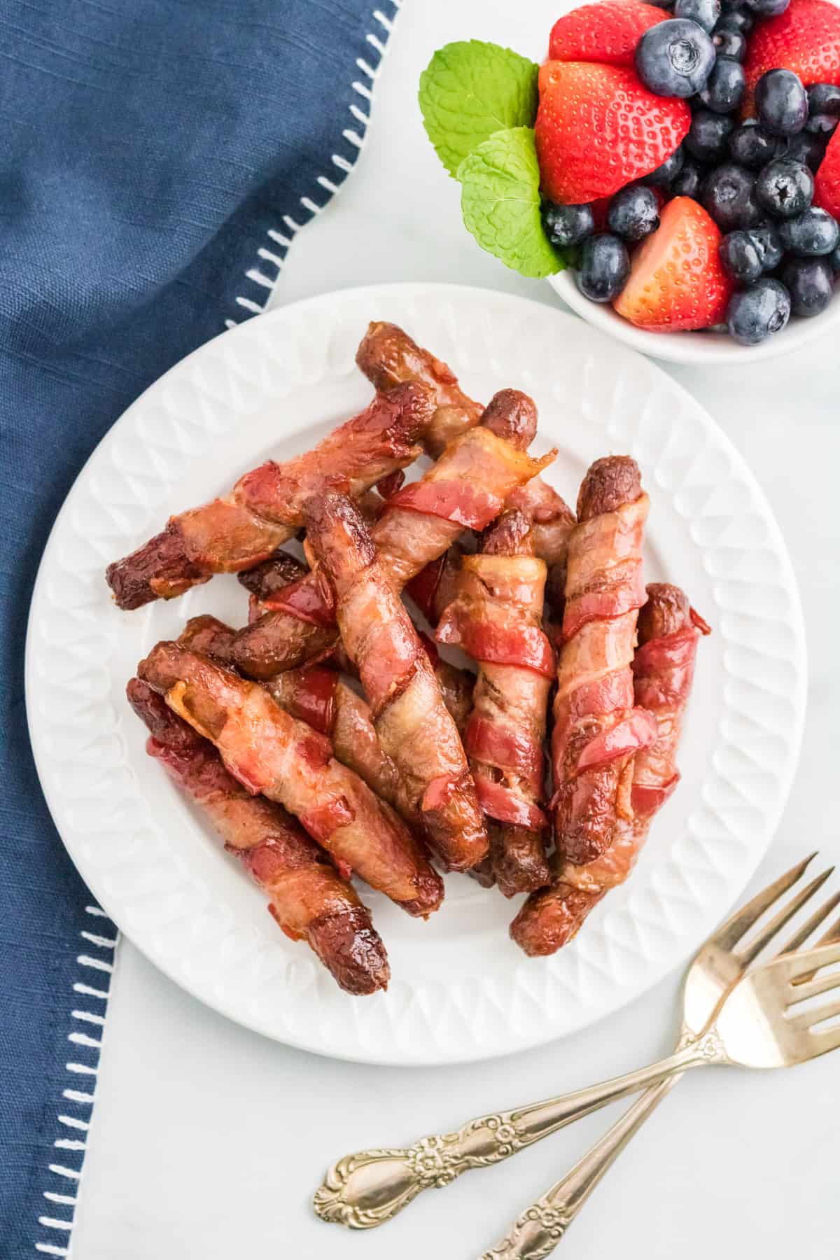 Overhead of stacked Bacon Wrapped Sausages on white plate with a bowl of fruit and forks beside it.