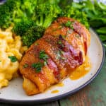 Square image of chicken breast on plate wrapped in bacon and topped with parsley. Served with macaroni and cheese and broccoli.