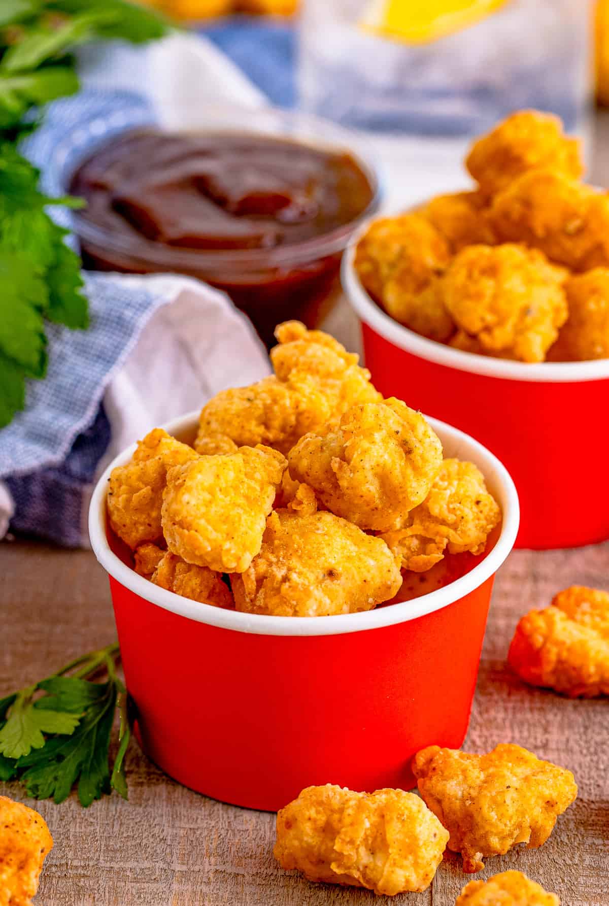 Popcorn Chicken Recipe in red cardboard cup with chicken pieces around it and BBQ sauce in background.