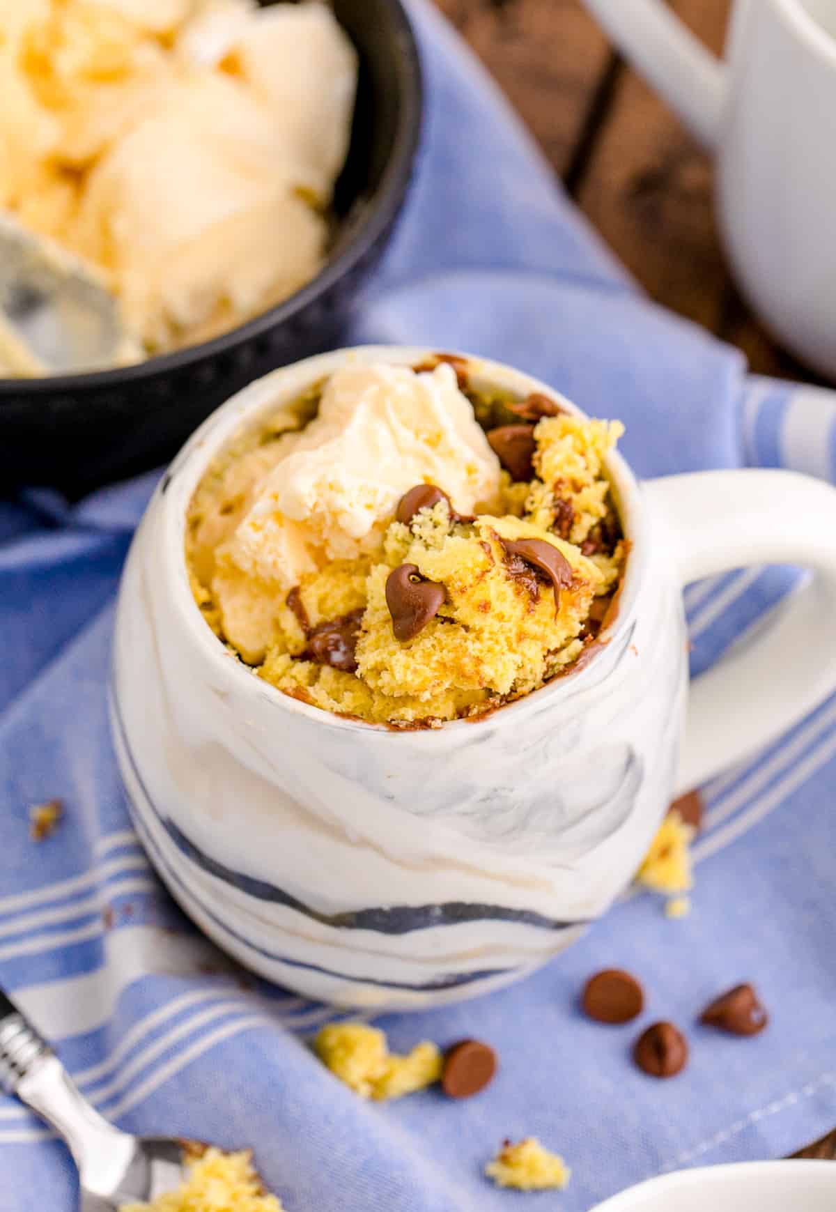 Chocolate Chip Mug Cake with ice cream added to mug on blue linen with ice cream in background surrounded by chocolate chips.