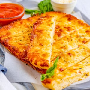 Square image of cut breadsticks with marinara in background and one stick laying on top of the other.