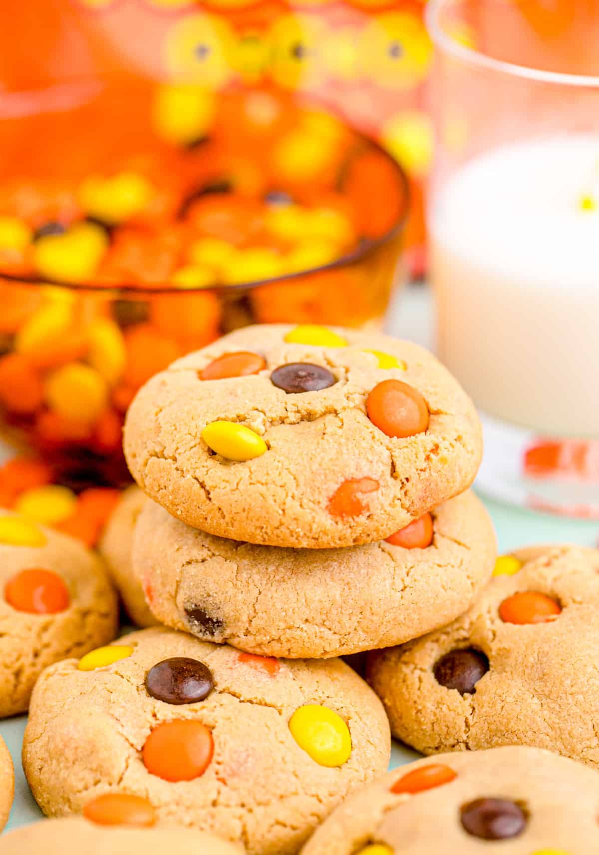 Stacked Reese's Peanut Butter Cookies with Reese's Pieces and milk in background.