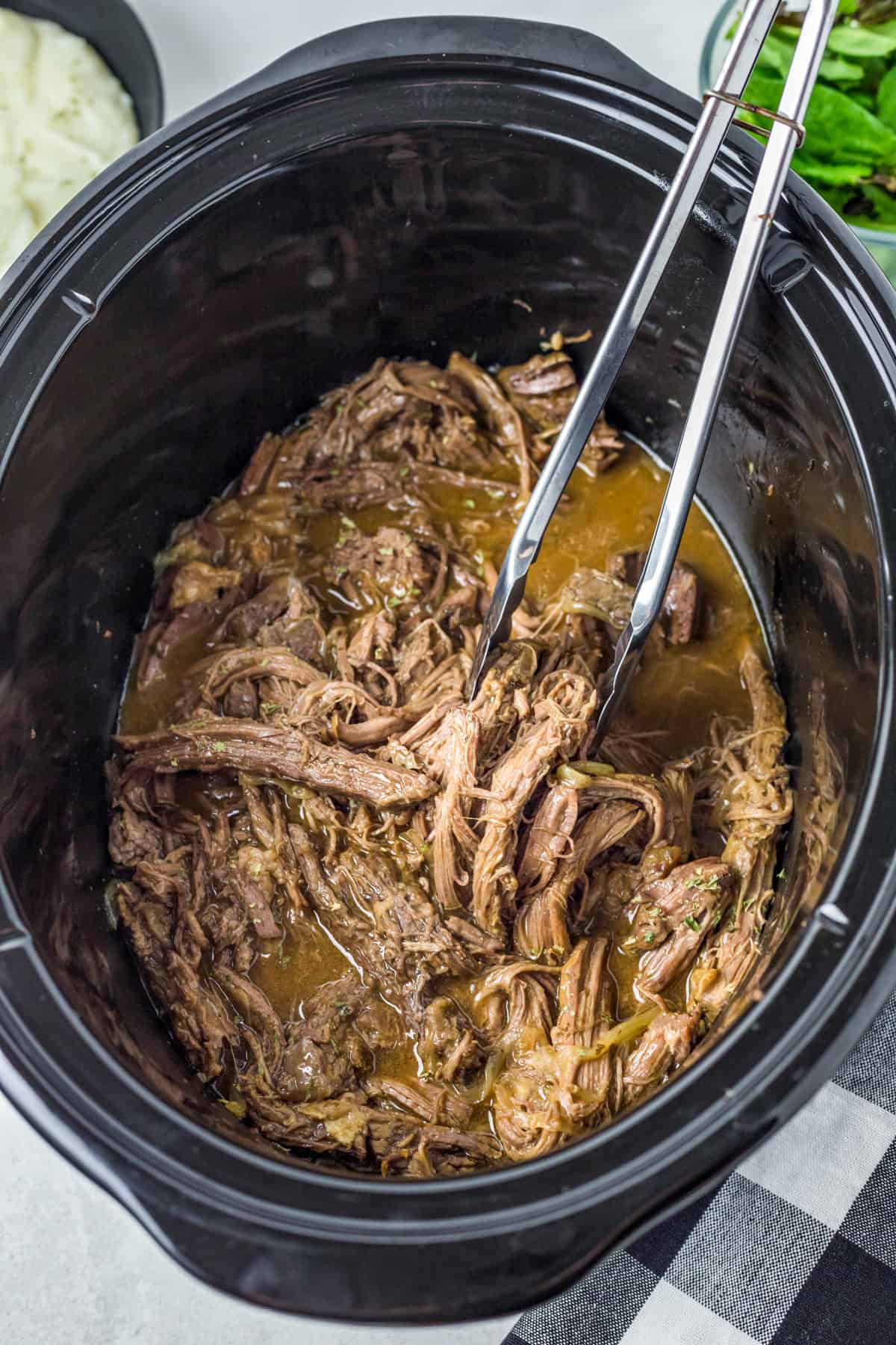 Finished Slow Cooker Shredded Beef in slow cooker with tongs holding some up.