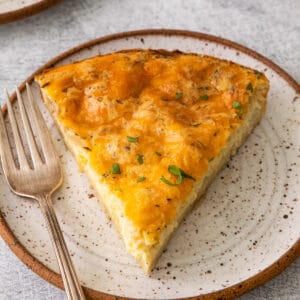 Close up square image of a slice of quiche on a white speckled plate with a fork.