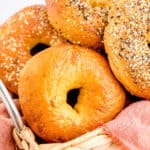 Close up square image of bagels in basket with pink linen and various toppings.