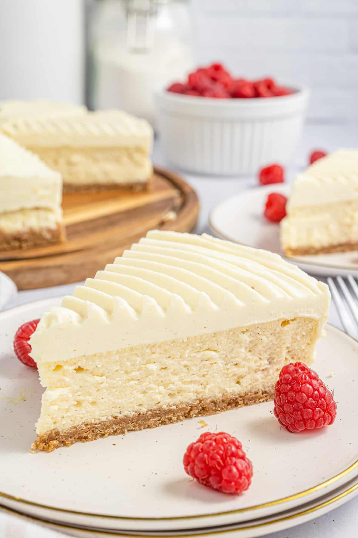 A slice of the Vanilla Bean Cheesecake on white plate with raspberries and full cheesecake in background.