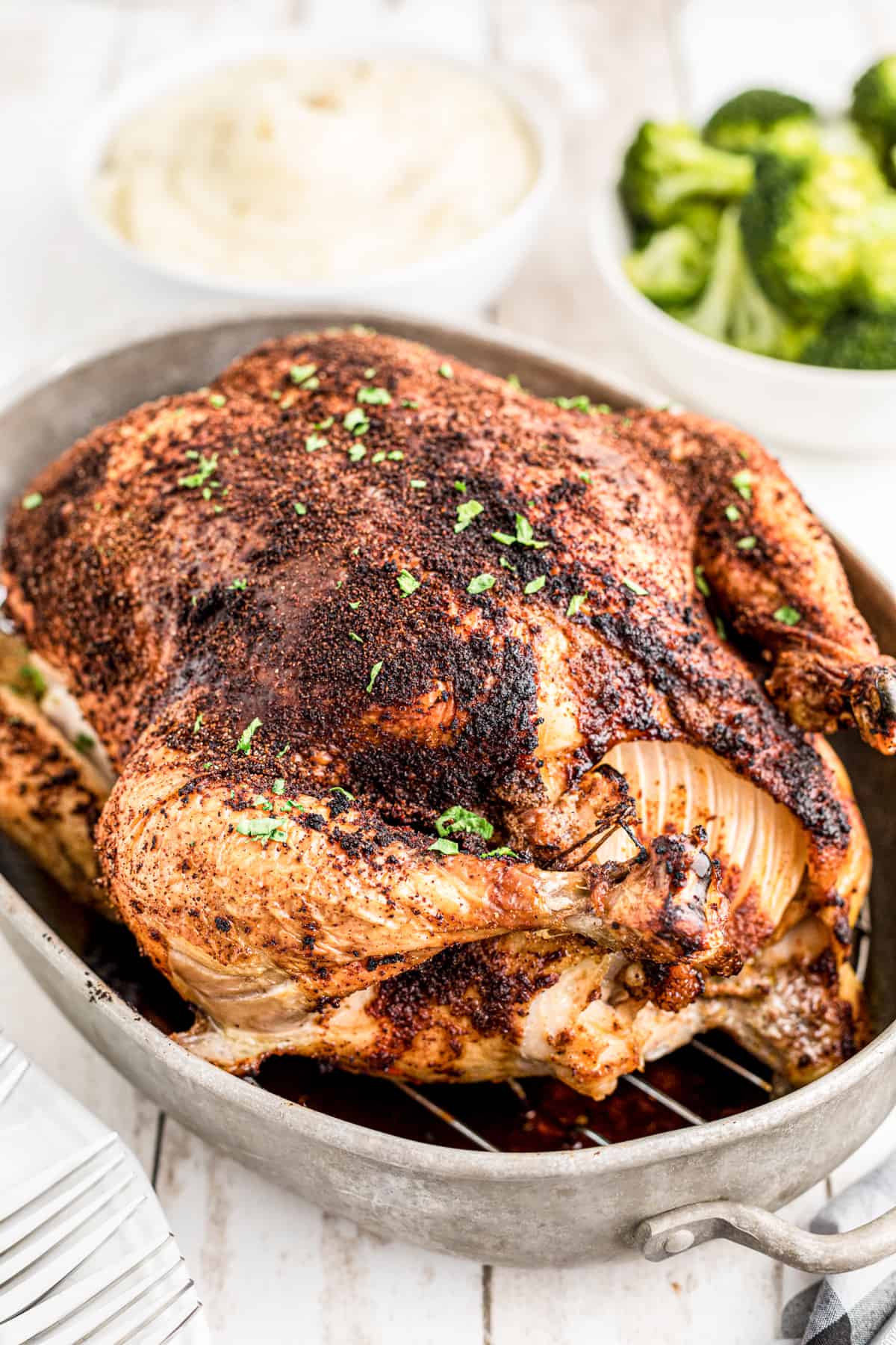 Whole Roasted Chicken finished in roasting pan with parsley and side dishes in background.