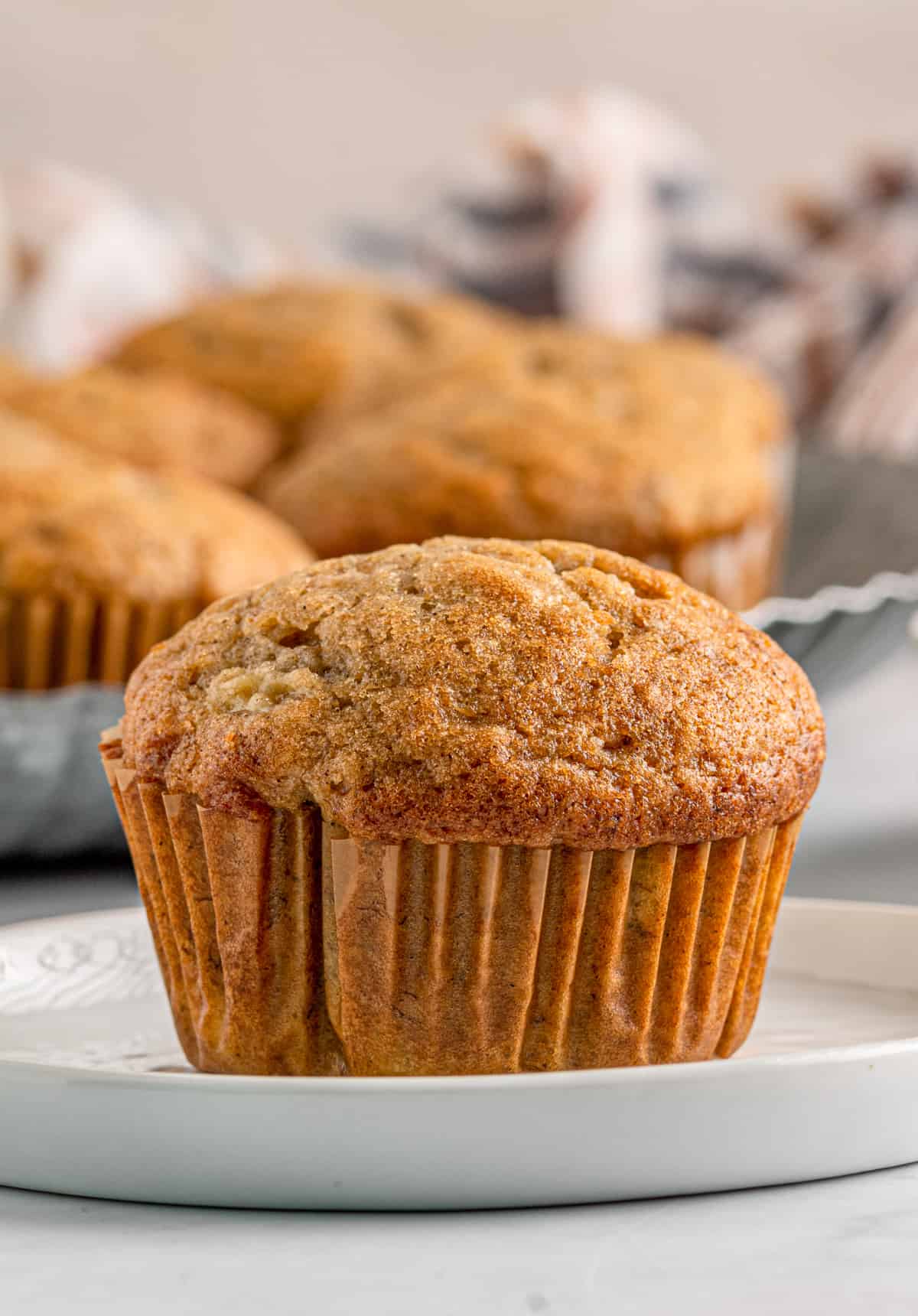 One of the Easy Banana Muffins on white plate with more muffins in background.