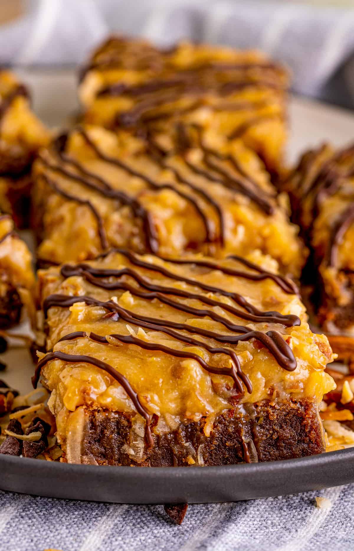 German Chocolate Brownies on black plate with pecan frosting and chocolate drizzle.