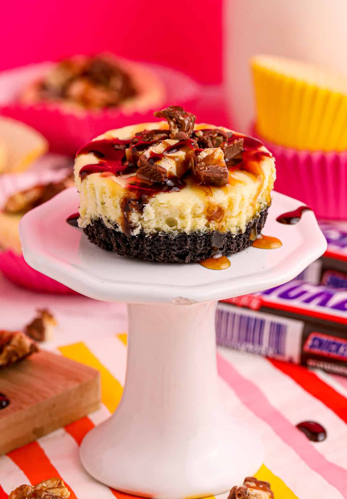 One of the Snickers Mini Cheesecakes on a cupcake stand with garnishes on top and dripping down the sides.