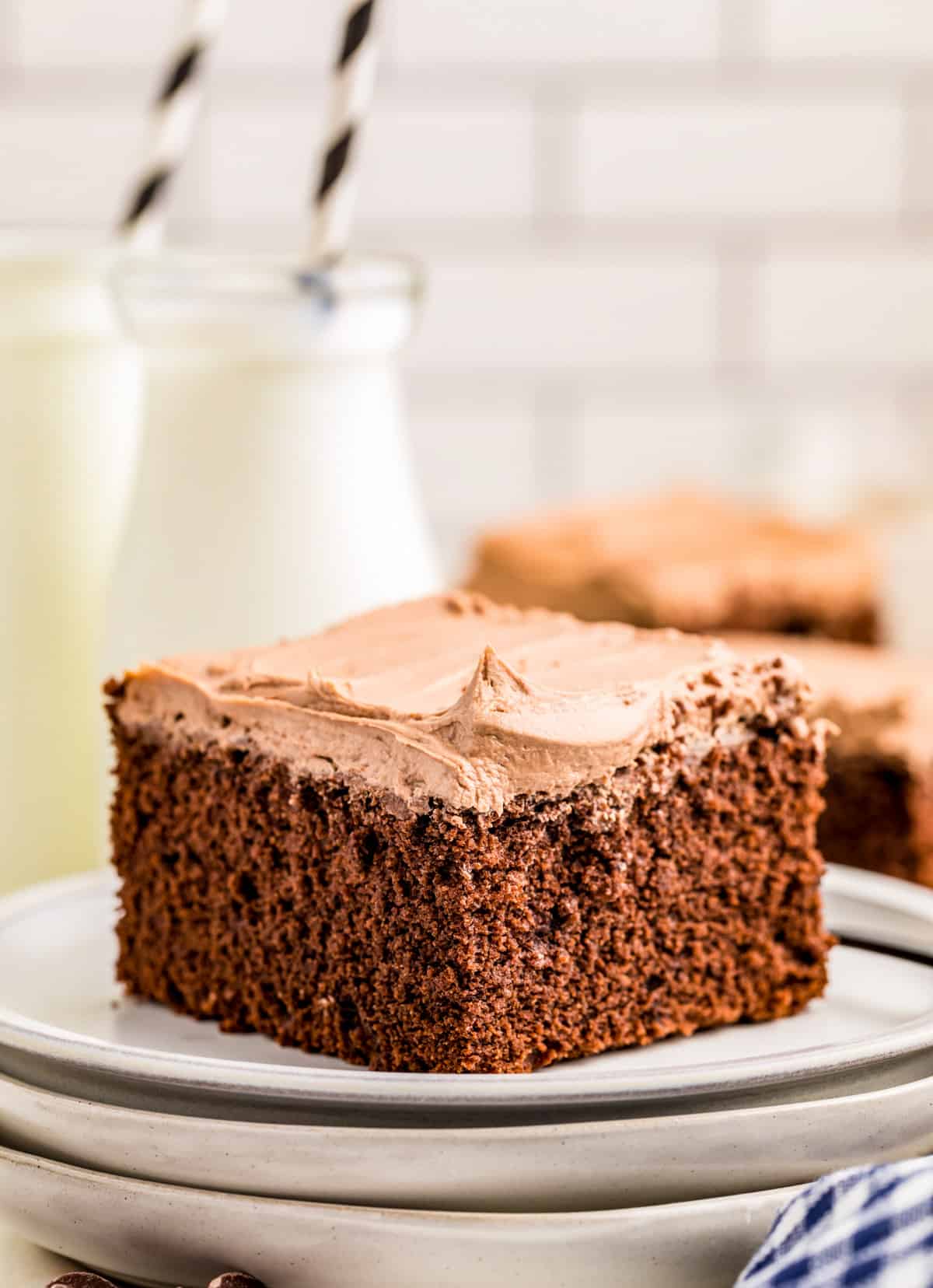 Slice of Easy Chocolate Cake on stacked plates with milk and cake slices in background.