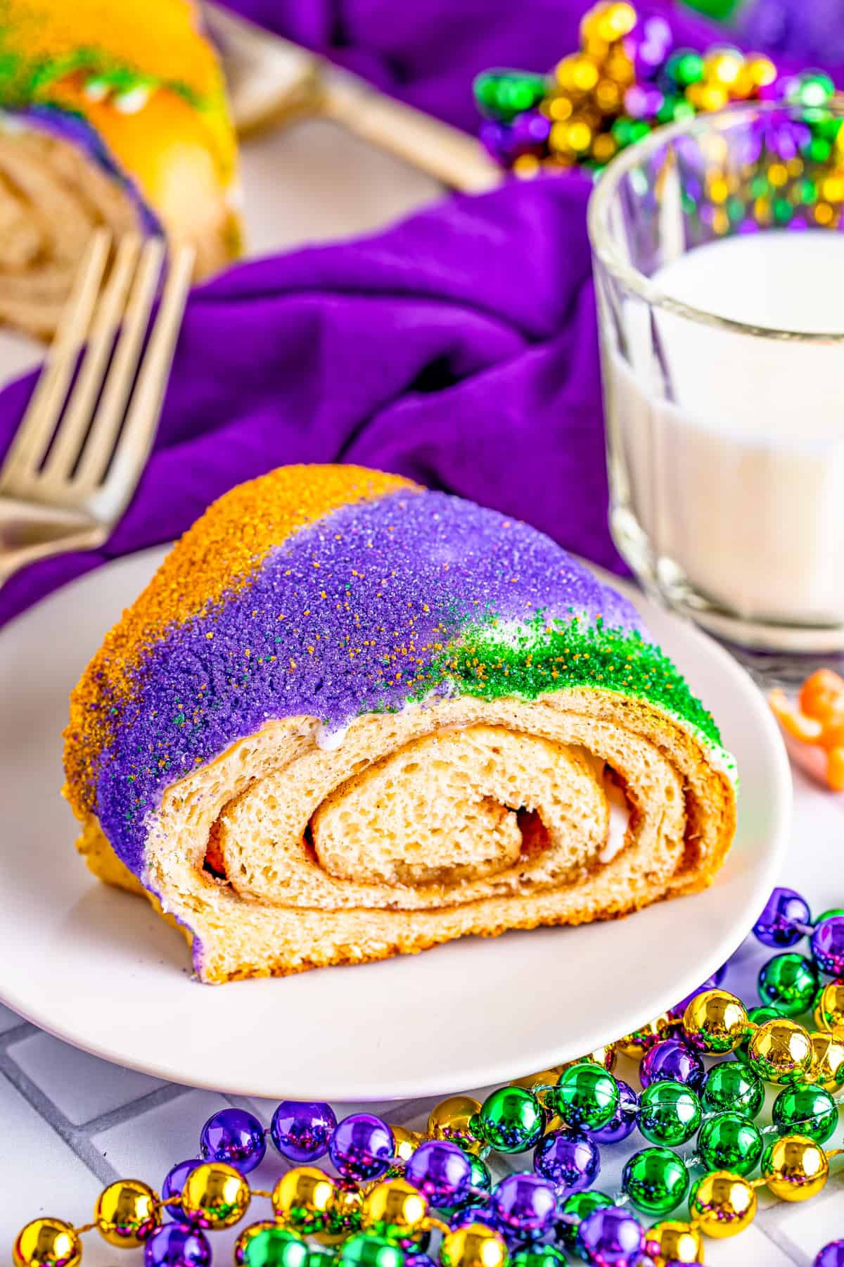 Slice of the King Cake Recipe on white plate showing topping and swirled inside surrounded by beads, milk and fork.