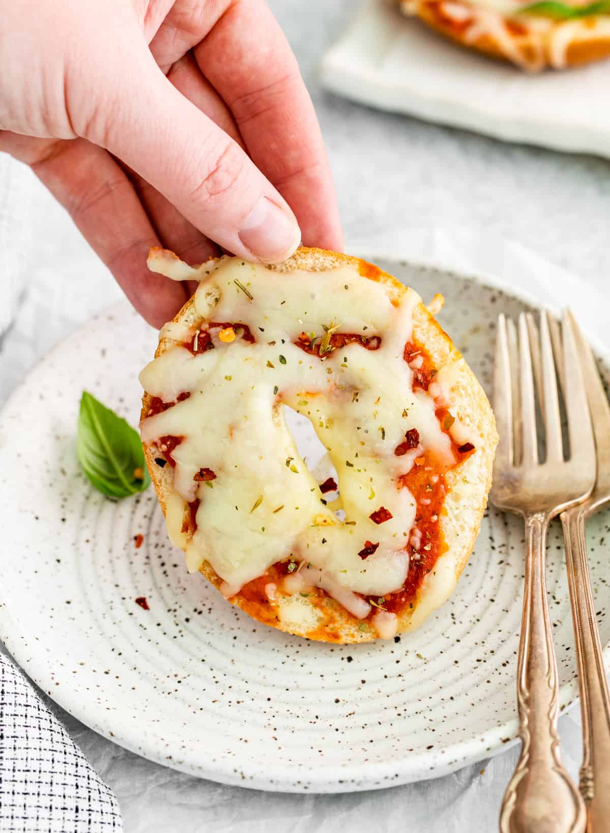 Hand holding up one of the Mini Pizza Bagels slightly off plate with forks and basil leaf.