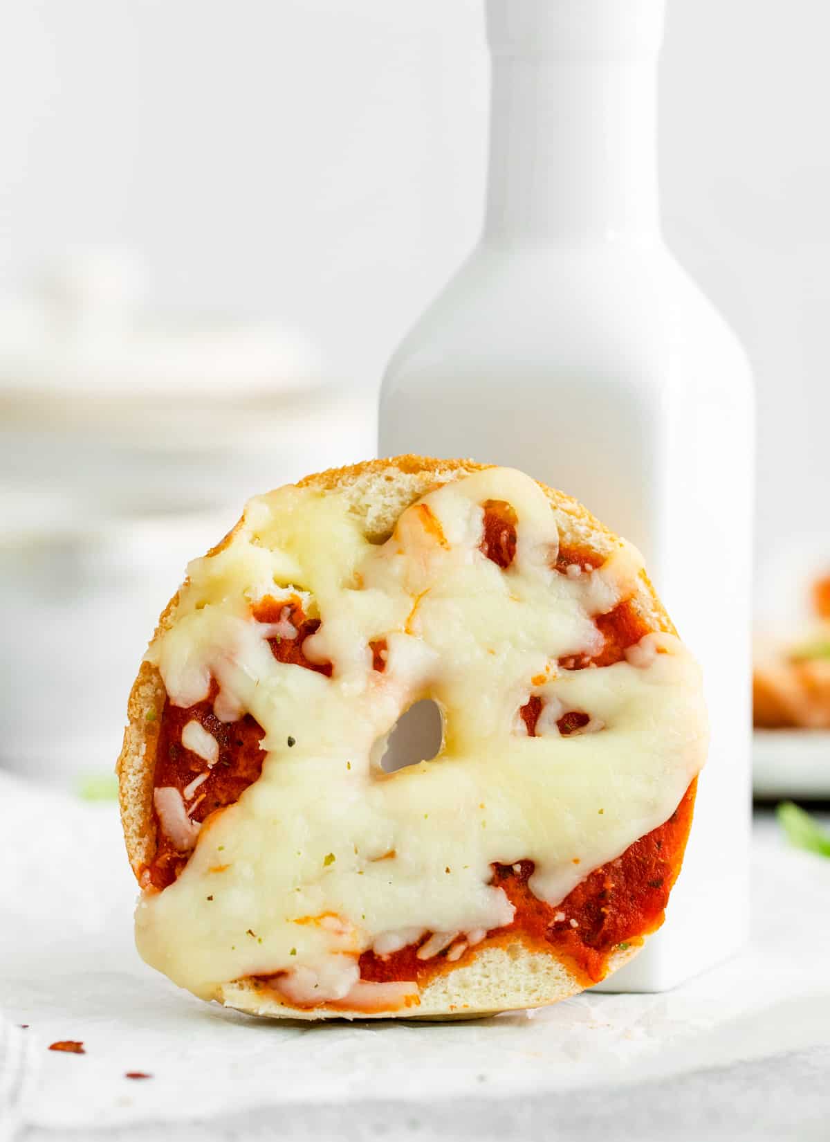 On of the finished Mini Pizza Bagels on parchment paper propped up on white container showing melted cheese.