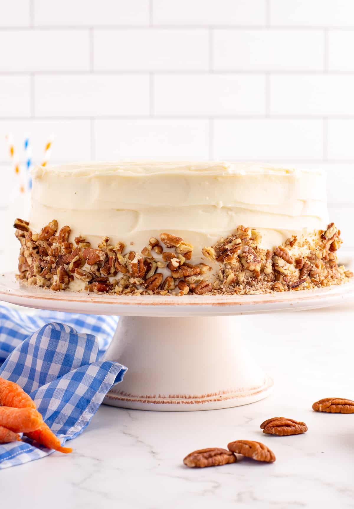 Finished Carrot Cake Recipe on white cake stand decorated with chopped nuts with nuts on the countertop beside cake stand.