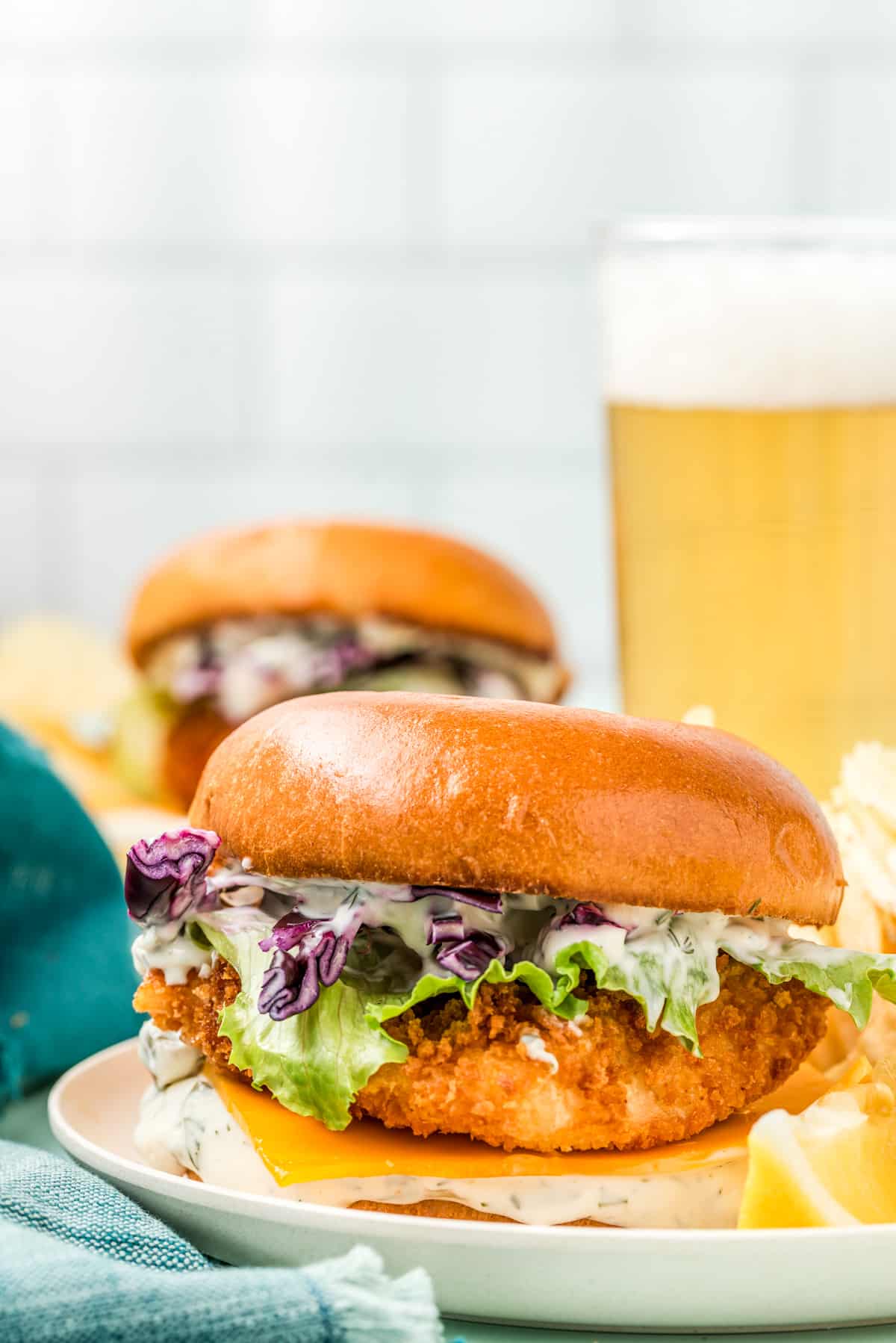 Crispy Fish Sandwich on plate with tartar sauce, cheese, lettuce and red cabbage with a beer in background.
