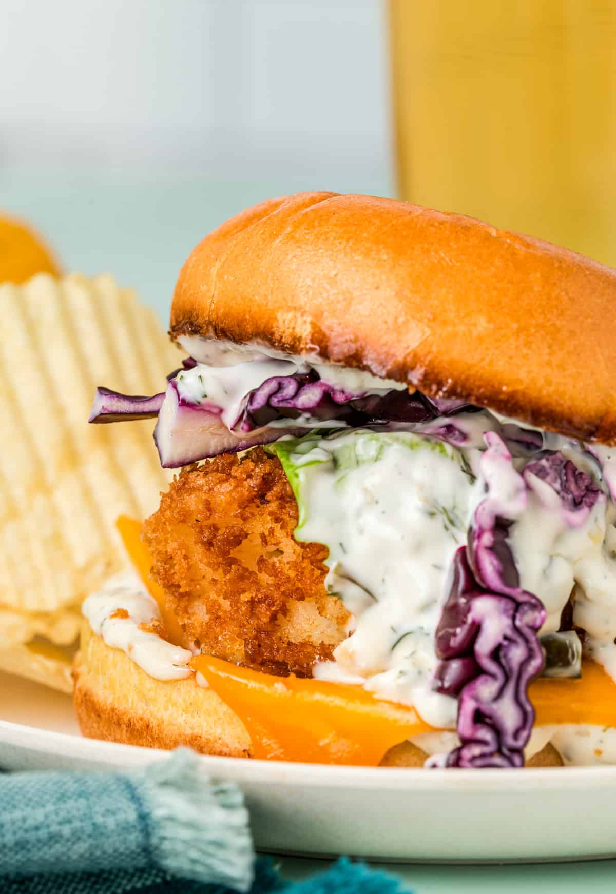Crispy Fish Sandwich on white plate with tartar sauce drizzling down side and potato chips on plate.