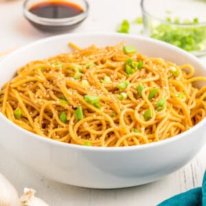 Close up square image of noodles in white bowl topped with green onions and white sesame seeds.