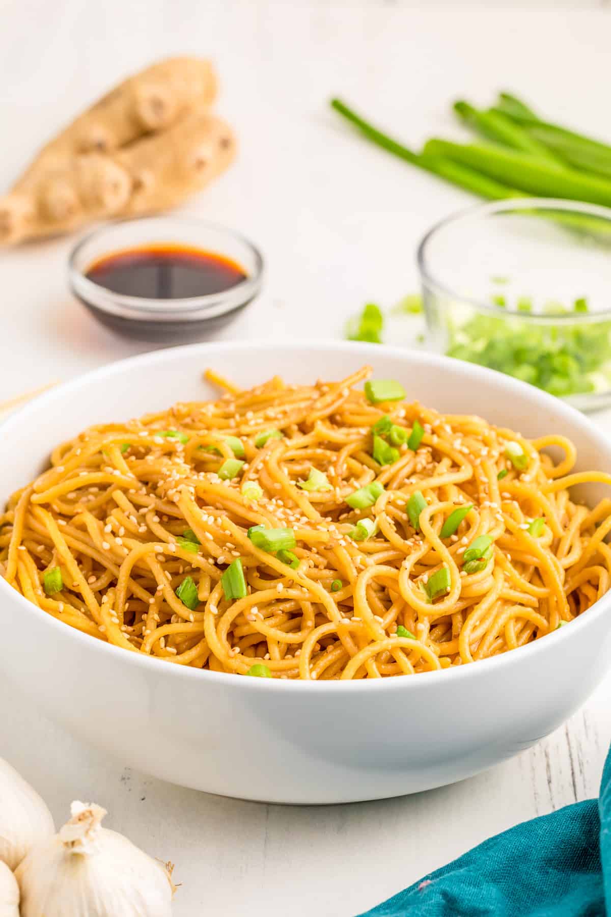 Hibachi Noodles served in a white bowl topped with green onions and white sesame seeds with other ingredients behind bowl.