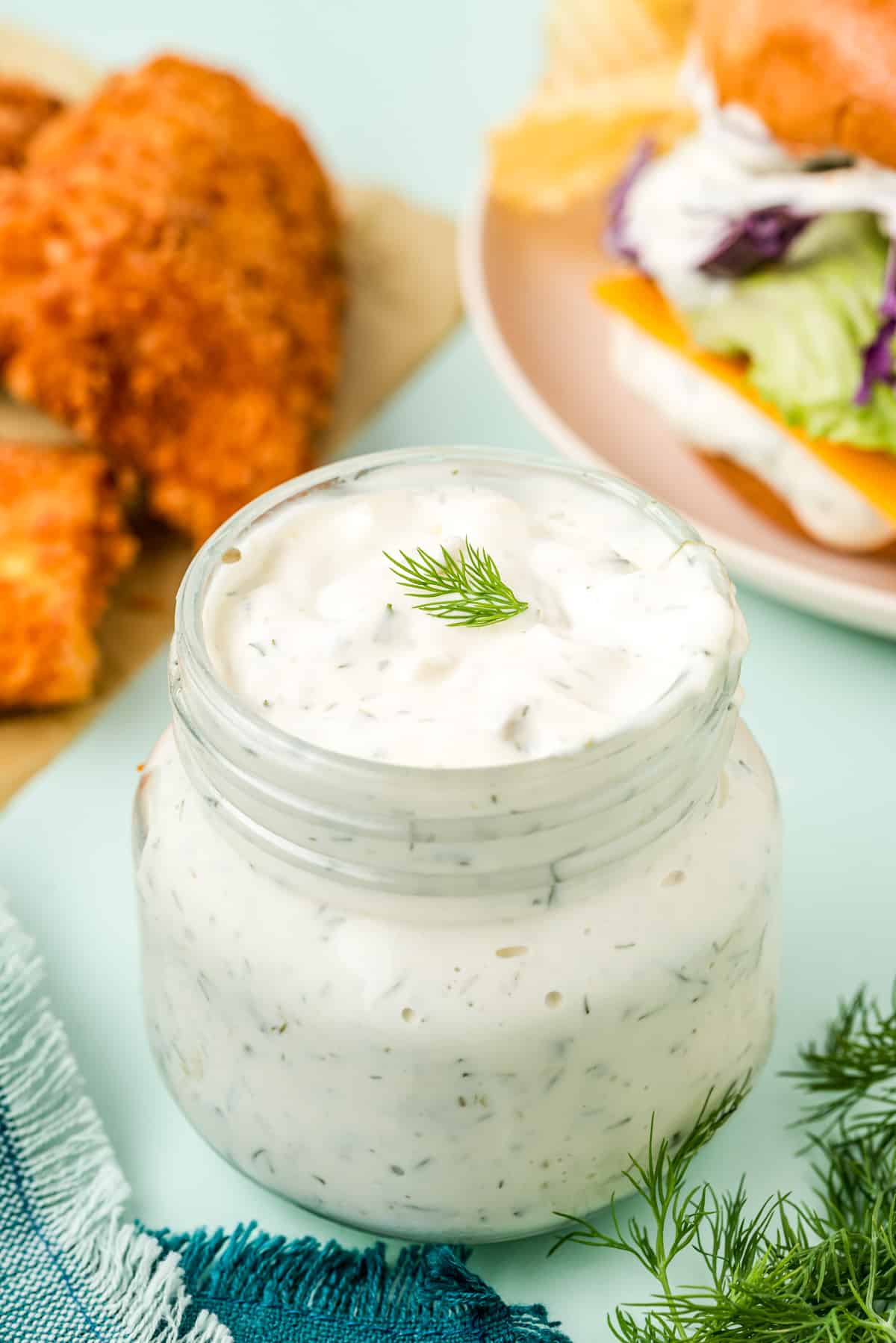 Homemade Tartar Sauce in glass jar with a sprig of dill and fish and fish sandwich in background.