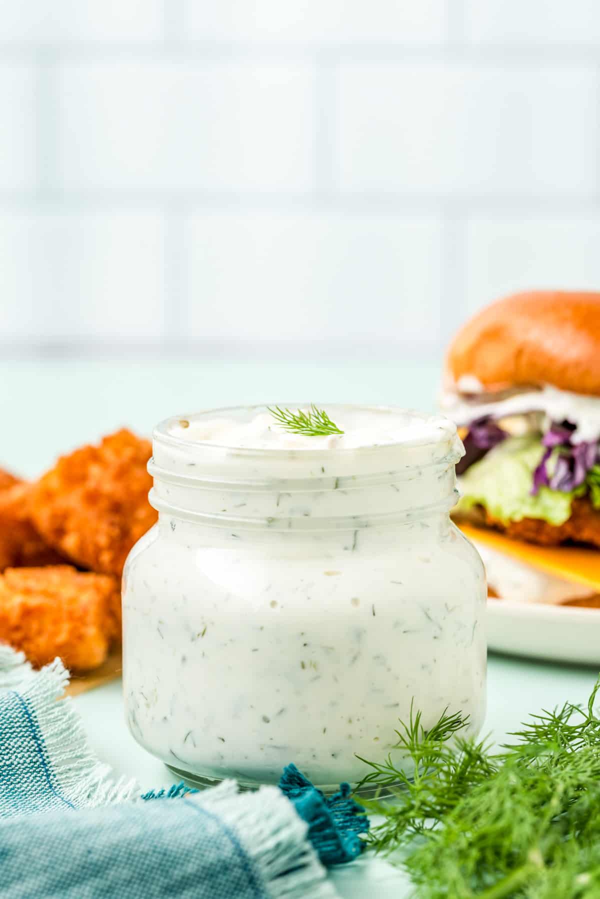Sauce in glass jar with fish and sandwich in background and dill in front of jar.