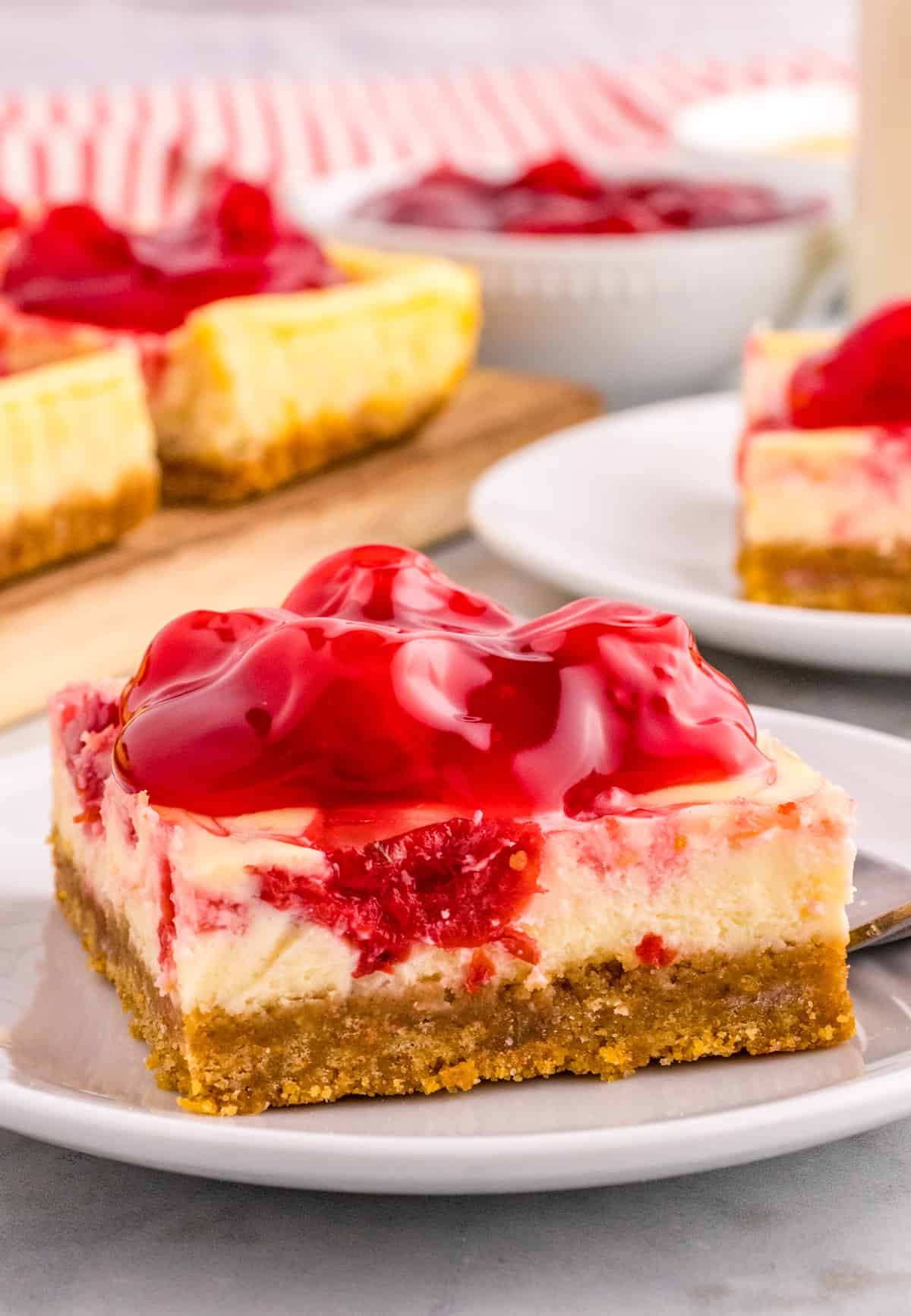 One of the Cherry Cheesecake Bars on a white plate topped with cherry pie filling with more bars in background.