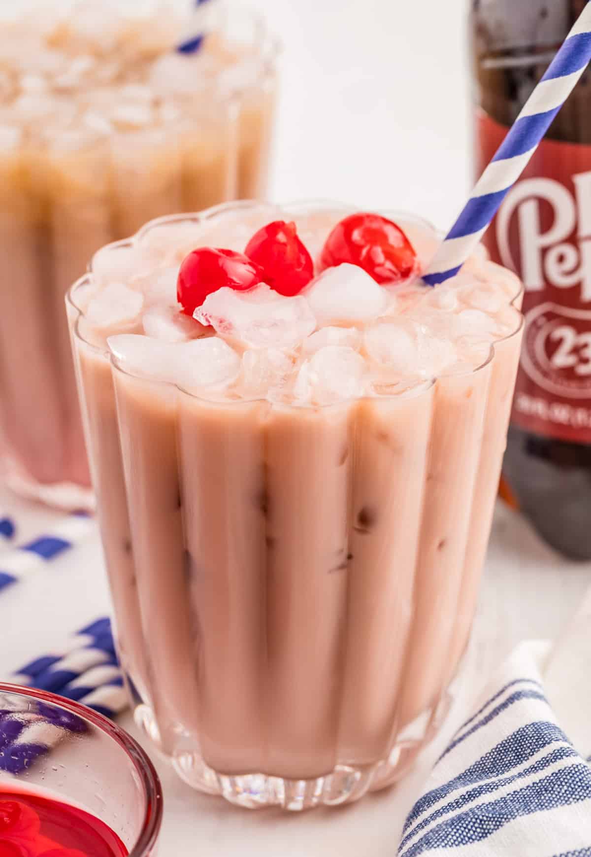 Dirty Soda in glass with ice, cherries and a blue and white straw with a bottle of doctor pepper in background.