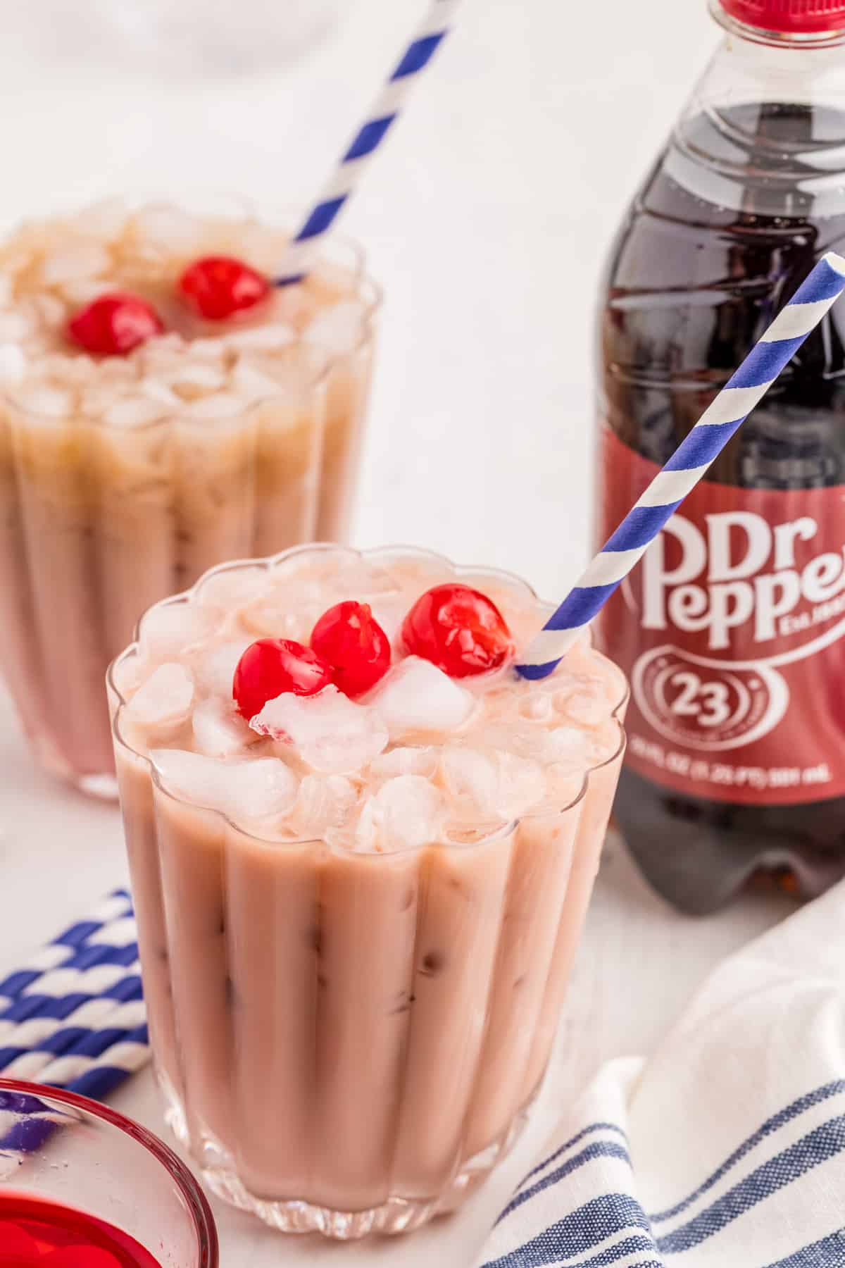 Two glasses of soda garnished with cherries and straws with a bottle of Dr. Pepper in background.
