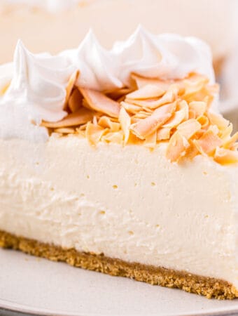 Close up square image of a slice of cheesecake on white plate topped with toasted coconut and whipped cream.